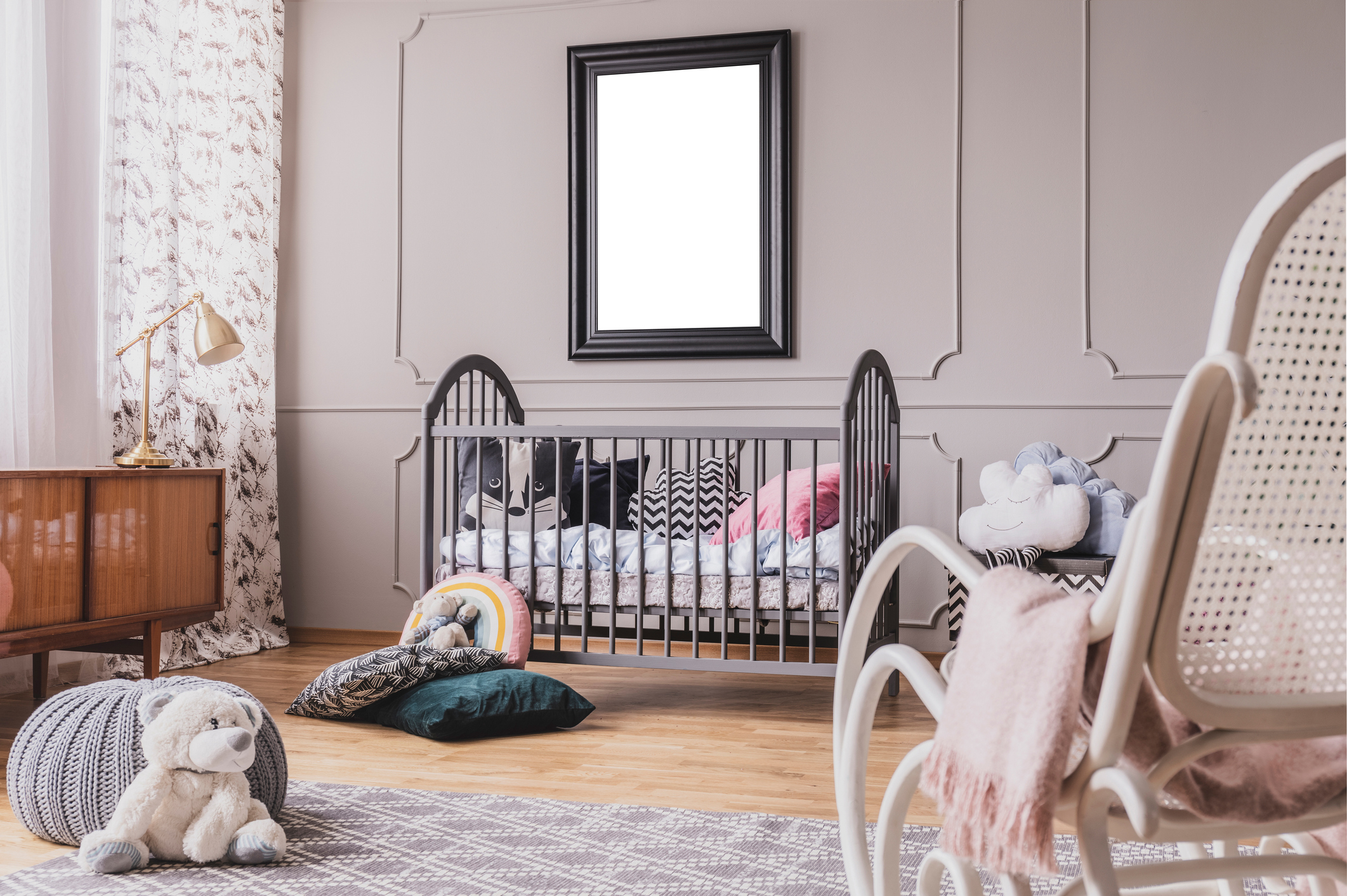 White teddy bear on the floor of stylish baby room interior with grey wooden crib with pillows, white rocking chair and mockup poster on the wall