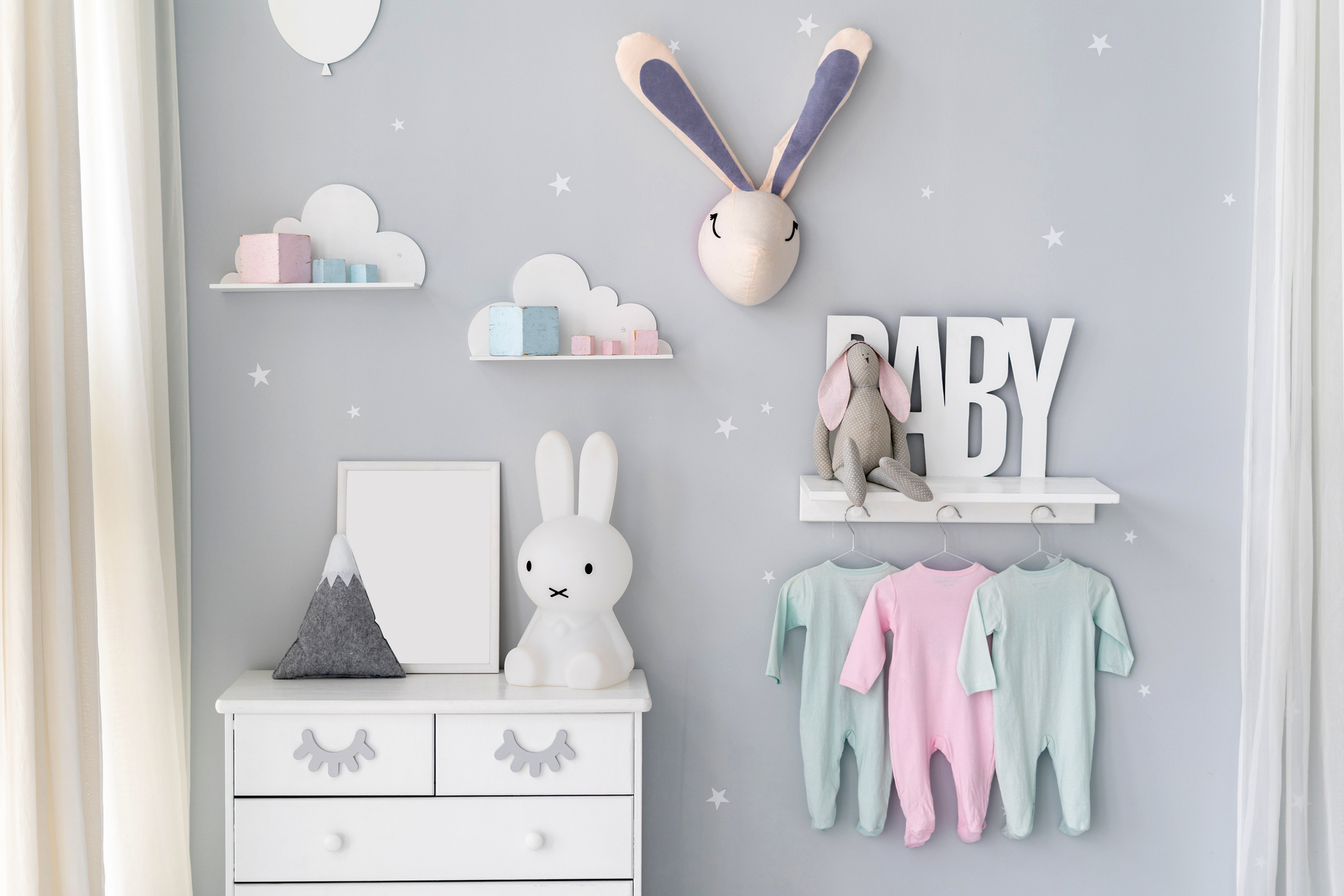 Light nursery room for newborn baby with commode, bunny toys, house decor and clothes wear on rack under shelf in nordic apartment interior style