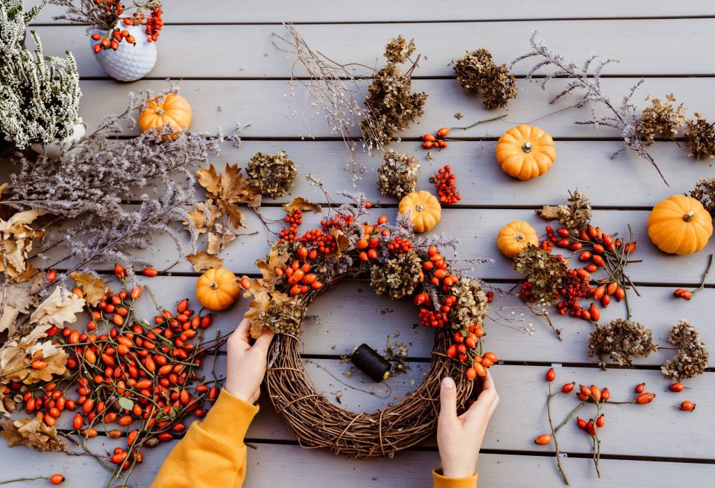 Girl making floral autumn door wreath using colorful rosehip berries, rowan, dry flowers and pumpkins. Fall flower decoration workshop, florist at work.