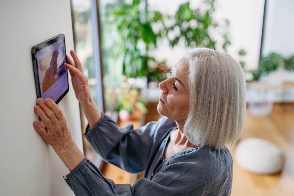 smart home, Mature woman adjusting smart thermostat, touch screen of smart home device. Older woman using smart technology at her home.